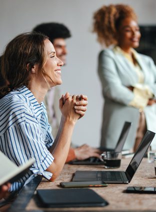 A side view of a smiling Caucasian entrepreneur using her laptop while attending a business meeting with a group of other employees.