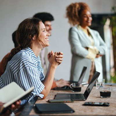 A side view of a smiling Caucasian entrepreneur using her laptop while attending a business meeting with a group of other employees.