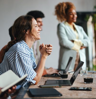 A side view of a smiling Caucasian entrepreneur using her laptop while attending a business meeting with a group of other employees.