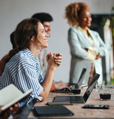 A side view of a smiling Caucasian entrepreneur using her laptop while attending a business meeting with a group of other employees.
