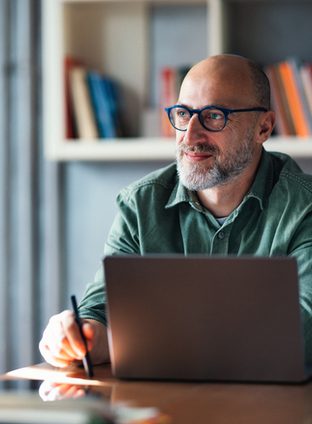 A thoughtful mature man working on his laptop in a modern office, surrounded by bookshelves, suggesting a productive and professional atmosphere ideal for work, creativity, or study.