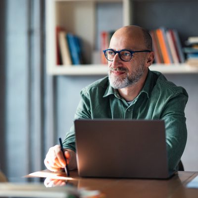 A thoughtful mature man working on his laptop in a modern office, surrounded by bookshelves, suggesting a productive and professional atmosphere ideal for work, creativity, or study.