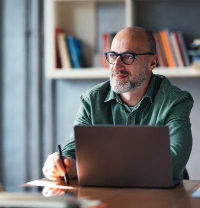 A thoughtful mature man working on his laptop in a modern office, surrounded by bookshelves, suggesting a productive and professional atmosphere ideal for work, creativity, or study.