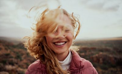 Portrait of young smiling woman face partially covered with flying hair standing at mountain - carefree woman