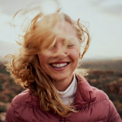 Portrait of young smiling woman face partially covered with flying hair standing at mountain - carefree woman