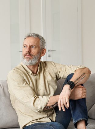 Relaxed mature older thoughtful tattooed hipster man relaxing sitting on sofa and thinking at home. Pensive calm stylish mid aged single bearded guy resting on couch at home looking away, reflecting.
