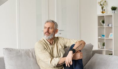 Relaxed mature older thoughtful tattooed hipster man relaxing sitting on sofa and thinking at home. Pensive calm stylish mid aged single bearded guy resting on couch at home looking away, reflecting.