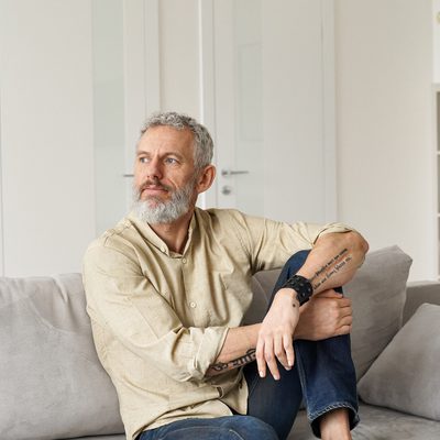 Relaxed mature older thoughtful tattooed hipster man relaxing sitting on sofa and thinking at home. Pensive calm stylish mid aged single bearded guy resting on couch at home looking away, reflecting.