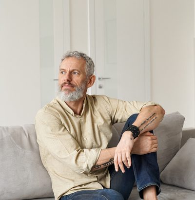 Relaxed mature older thoughtful tattooed hipster man relaxing sitting on sofa and thinking at home. Pensive calm stylish mid aged single bearded guy resting on couch at home looking away, reflecting.