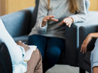 An unrecognizable mature adult woman gestures while talking to the unrecognizable mid adult male therapist. Her unrecognizable young adult daughter sits quietly nearby.