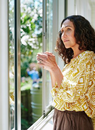 Content mature woman drinking coffee while looking out at the view from a window in her living room at home