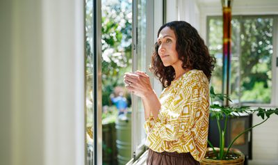 Content mature woman drinking coffee while looking out at the view from a window in her living room at home