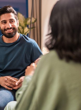 A cheerful young Arabic couple enjoys quality time together in a modern home, chatting comfortably on a beige sofa, highlighted by soft indoor lighting and contemporary decor.