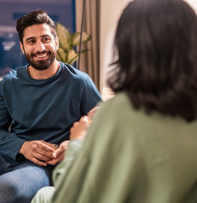 A cheerful young Arabic couple enjoys quality time together in a modern home, chatting comfortably on a beige sofa, highlighted by soft indoor lighting and contemporary decor.