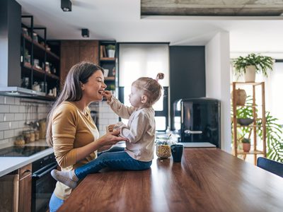 Mother and toddler sharing a joyful moment in a modern kitchen, with the child feeding the mother while both smile amidst sleek appliances and indoor plants