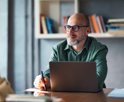 A thoughtful mature man working on his laptop in a modern office, surrounded by bookshelves, suggesting a productive and professional atmosphere ideal for work, creativity, or study.