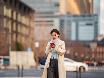 A young Japanese woman in a beige trench coat walks through an open plaza near Tokyo Station, focused on her smartphone with a handbag over her shoulder. The iconic red brick station building and modern skyscrapers frame the scene, capturing urban lifestyle and digital connectivity in central Tokyo.
