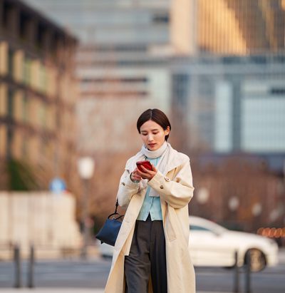 A young Japanese woman in a beige trench coat walks through an open plaza near Tokyo Station, focused on her smartphone with a handbag over her shoulder. The iconic red brick station building and modern skyscrapers frame the scene, capturing urban lifestyle and digital connectivity in central Tokyo.