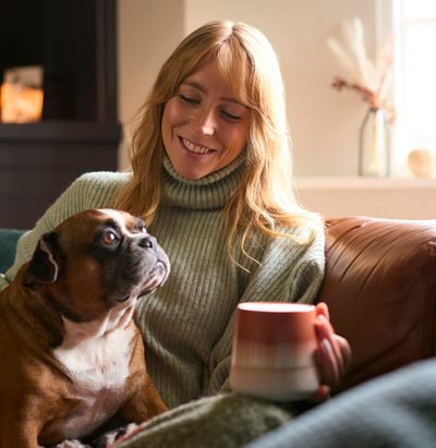 Woman At Home In Winter Jumper With Hot Drink Of Tea Or Coffee In Cup Stroking Pet French Bulldog