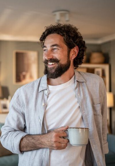 Happy man with beard and curly hair holding a mug and smiling while looking away in his cozy living room, enjoying a moment of peace at home
