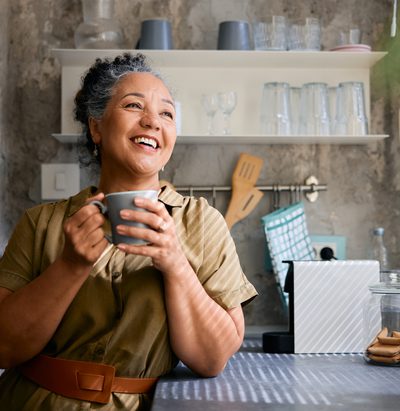 Cheerful mature woman sipping coffee in a modern kitchen, smiling while gazing out the window, embracing the peaceful morning and enjoying a moment of tranquility and relaxation