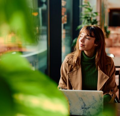 Woman freelancer taking a break from work, sitting at a cafe table with her laptop, contemplating while looking away with sunlight streaming through the window
