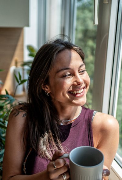 Cheerful young woman is holding a mug and looking out the window in her modern and bright apartment, enjoying the morning sunlight and the view of the trees outside