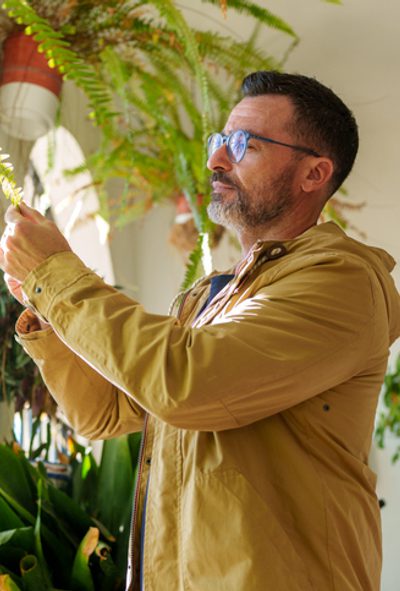 Mature adult man checking a fern leaf, enjoying his gardening hobby on a sunny day at a patio filled with plants