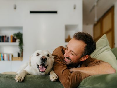 Photo of a man cuddling with his dog in the living room of their house.