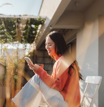 Photo of a smiling Japanese woman using a smartphone while sipping coffee on the balcony of her loft apartment